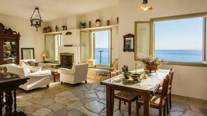 A spacious living room with a stone floor, a fireplace, and a large window overlooking the sea. - ReGenesis Villas (Tinos)