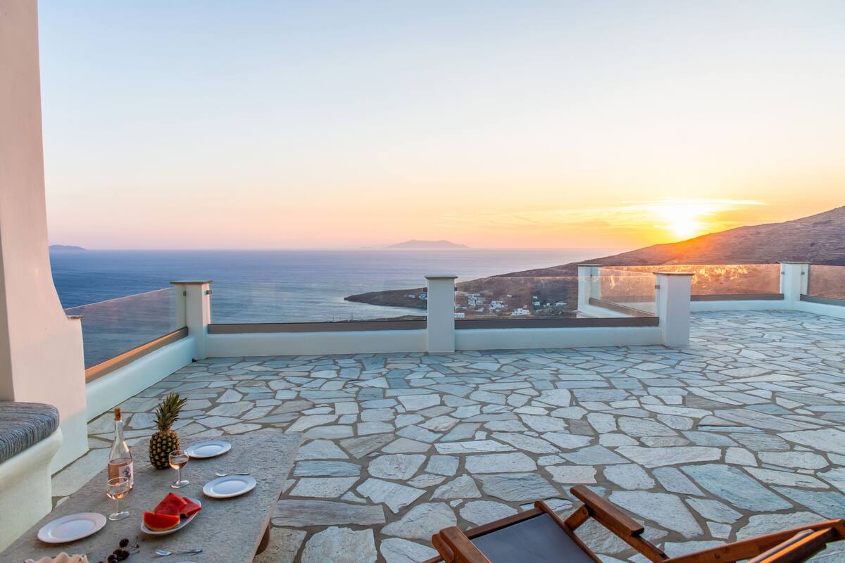 Una terraza con suelo de piedra, una mesa lista para dos personas y vistas al mar al atardecer.