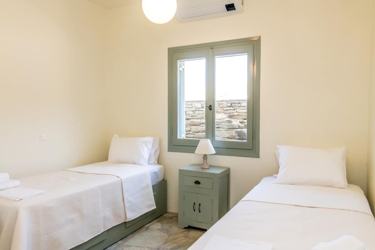 Una habitación con dos camas, una mesita de noche con lámpara y una ventana con vista a muros de piedra.