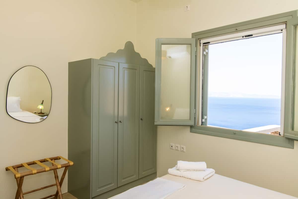 Un dormitorio con cama, armario, espejo, ventana con vista al mar y una mesita.