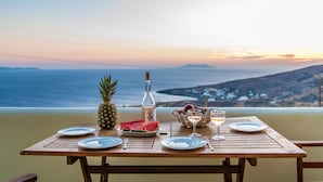 A wooden table set with plates, a pineapple, watermelon slices, a bottle, and a glass of white wine, overlooking a coastal view. - ReGenesis Villas (Tinos)