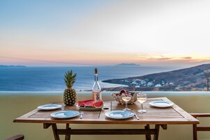 Una mesa de madera dispuesta con platos, una piña, rebanadas de sandía, una botella y una copa de vino blanco, con vistas a la costa.