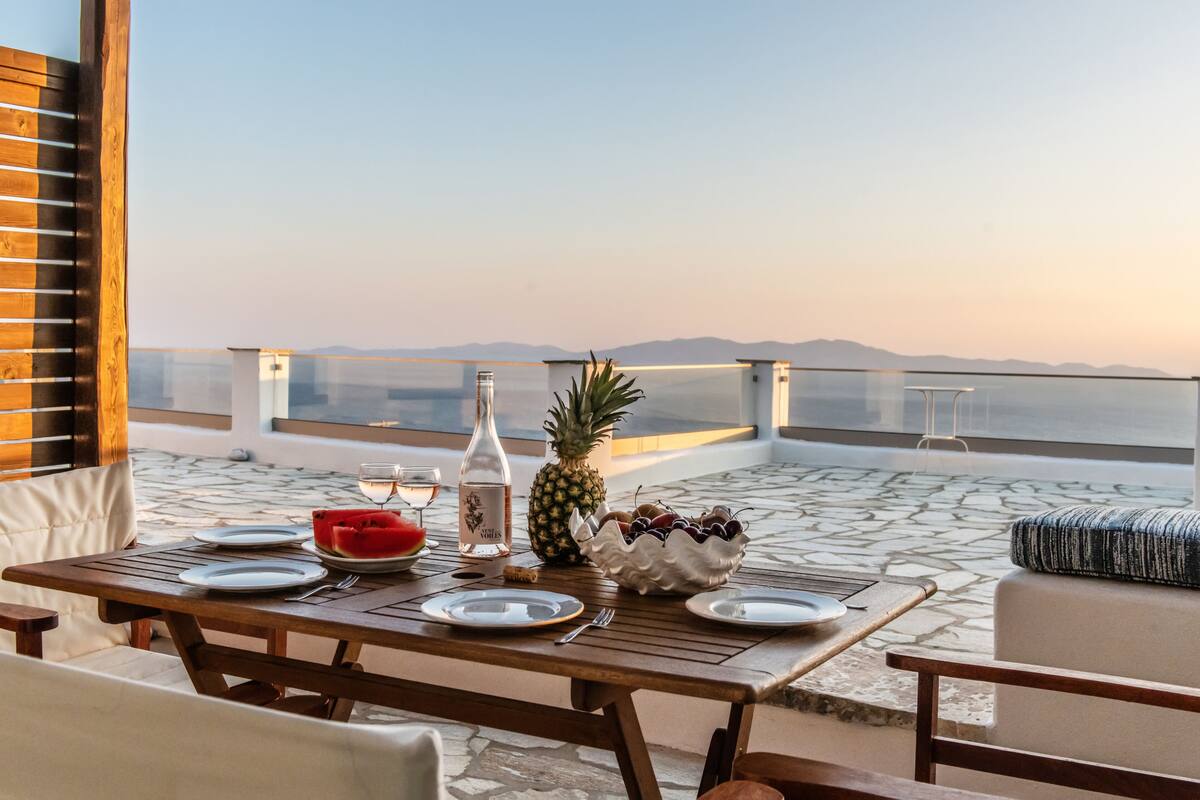 Una mesa de madera dispuesta con platos, una botella, una piña y un bol de uvas en un balcón con vistas despejadas a las montañas.