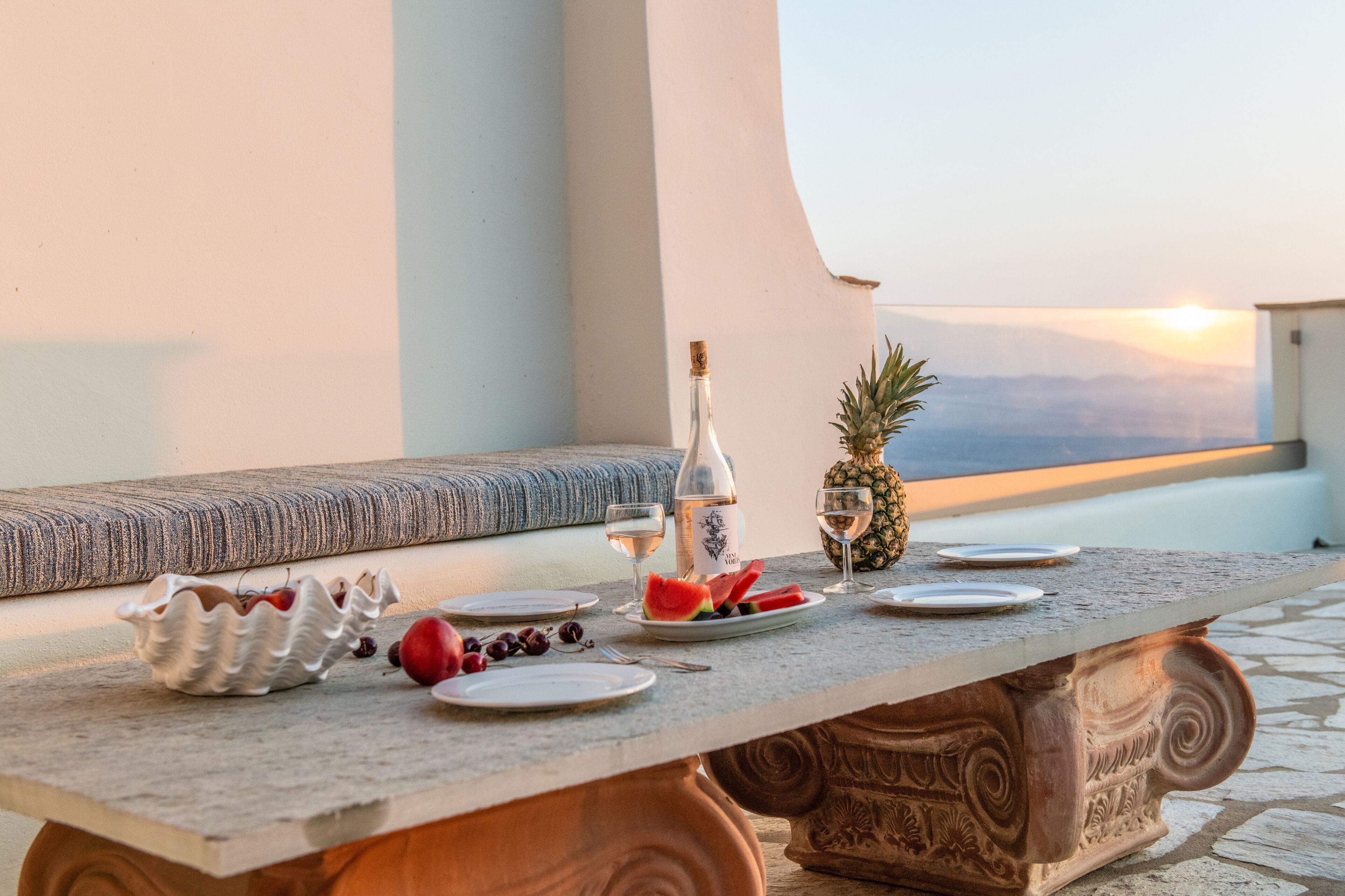 Een tafel gedekt met borden, een fles en fruit op een balkon met uitzicht op de zonsondergang.