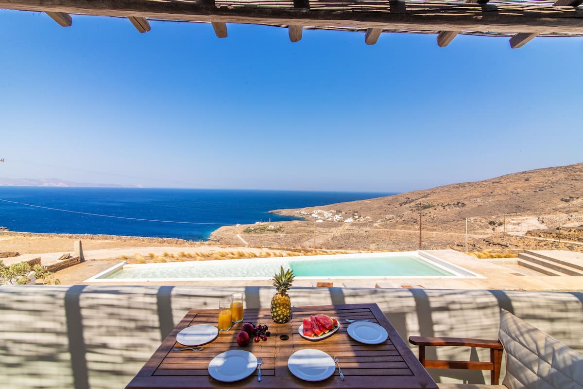 Una mesa de comedor con platos, cubiertos y fruta, con vistas a una piscina y un paisaje pintoresco del mar y las colinas.