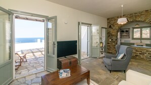 A living room with a stone fireplace, a television, and a view of the sea through open doors. - ReGenesis Villas (Tinos)