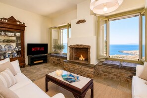A living room with a stone fireplace, a television, and a view of the sea. - ReGenesis Villas (Tinos)