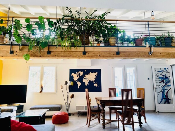 A spacious living area with a high ceiling, wooden beams, a dining table with chairs, a red ottoman, a television, and a world map on the wall.
