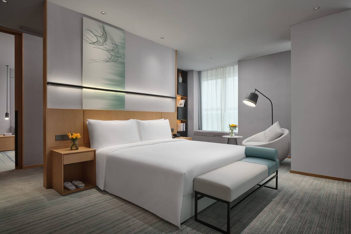 A modern hotel room with a large bed, bedside table, lamp, and a view of the city.