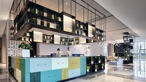 A modern reception area with a multi-tiered shelving unit displaying bottles, a colorful counter with drawers, and a large window offering a view of the outdoors. - Q-Box Hotel Shangqiu Expo Center (Shangqiu)
