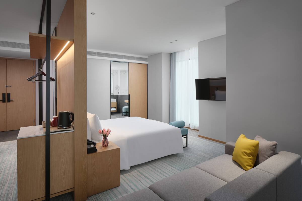 A modern hotel room with a large bed, a sofa, a desk with a coffee maker, and a TV mounted on the wall.