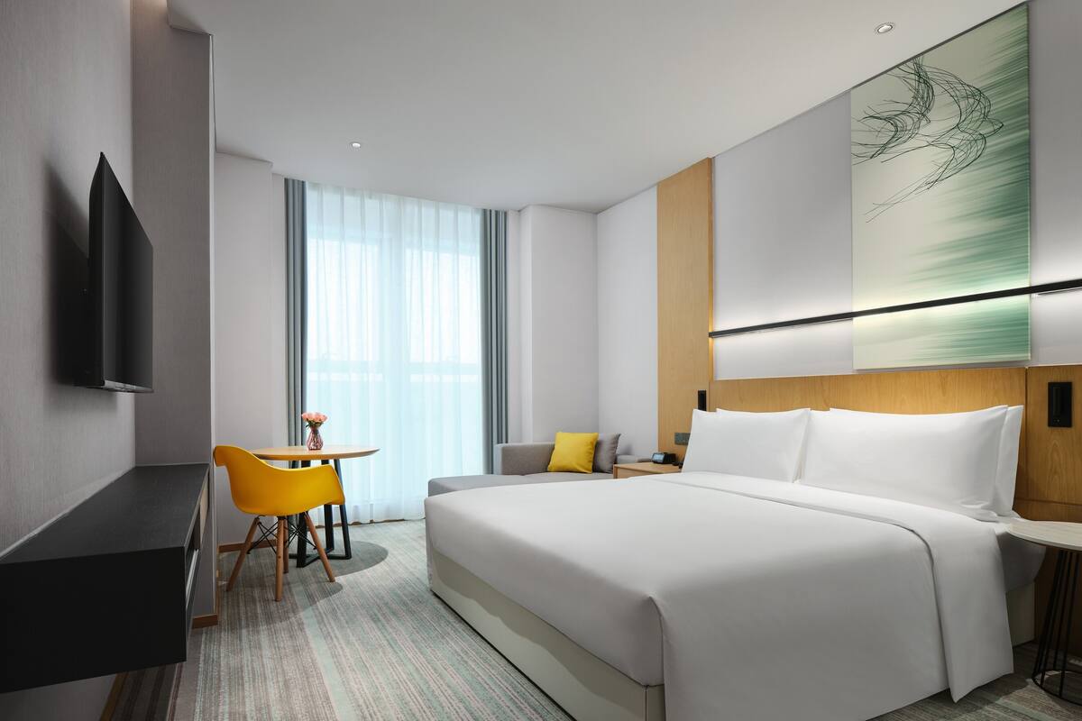 A modern hotel room with a large bed, a desk, a chair, and a TV mounted on the wall.