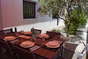 Mesa de madera para comer con platos, vasos y cubiertos, ubicada al aire libre con un árbol y un edificio de fondo.
