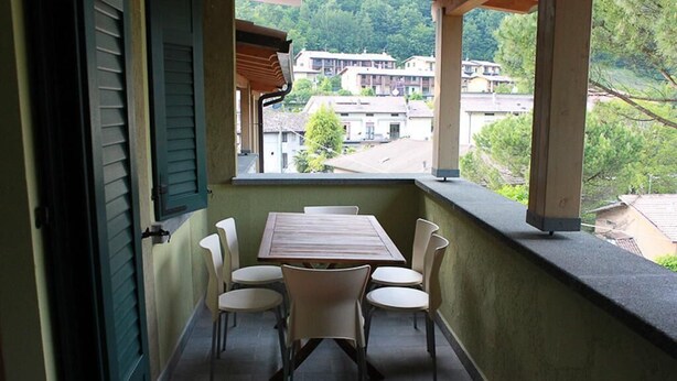 A balcony with a table and chairs, a view of buildings in the background, and a door with green shutters. - Two Bedroom Apartment (IDRO)