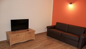 A room with a wooden cabinet, a flat-screen TV, and a brown sofa against a white wall with an orange accent wall. - Two Bedroom Apartment (IDRO)