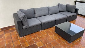 Un sofá gris trenzado con cojines y una mesa de centro de vidrio negro en un patio con baldosas.