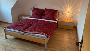 Un dormitorio con techo inclinado, una cama con manta roja, dos mesitas de noche y una lámpara fijada a la pared.