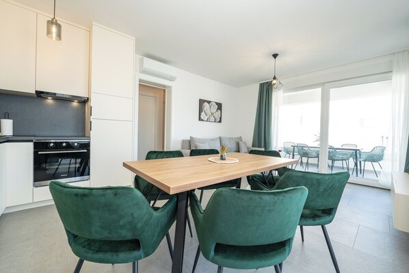 A modern kitchen with a built-in oven, a dining table with four green chairs, and a large window with a view of outdoor chairs.