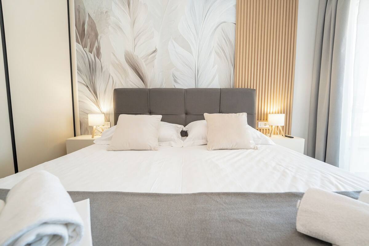 Una cama bien tendida con sábanas blancas y dos almohadas, cabecera gris y una gran obra de arte abstracto en la pared.