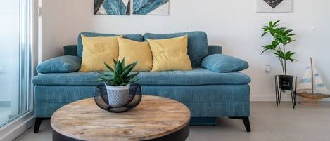 Un salon moderne avec un canapé de couleur turquoise, des coussins jaunes, une table basse ronde en bois et des œuvres d’art abstraites accrochées aux murs.