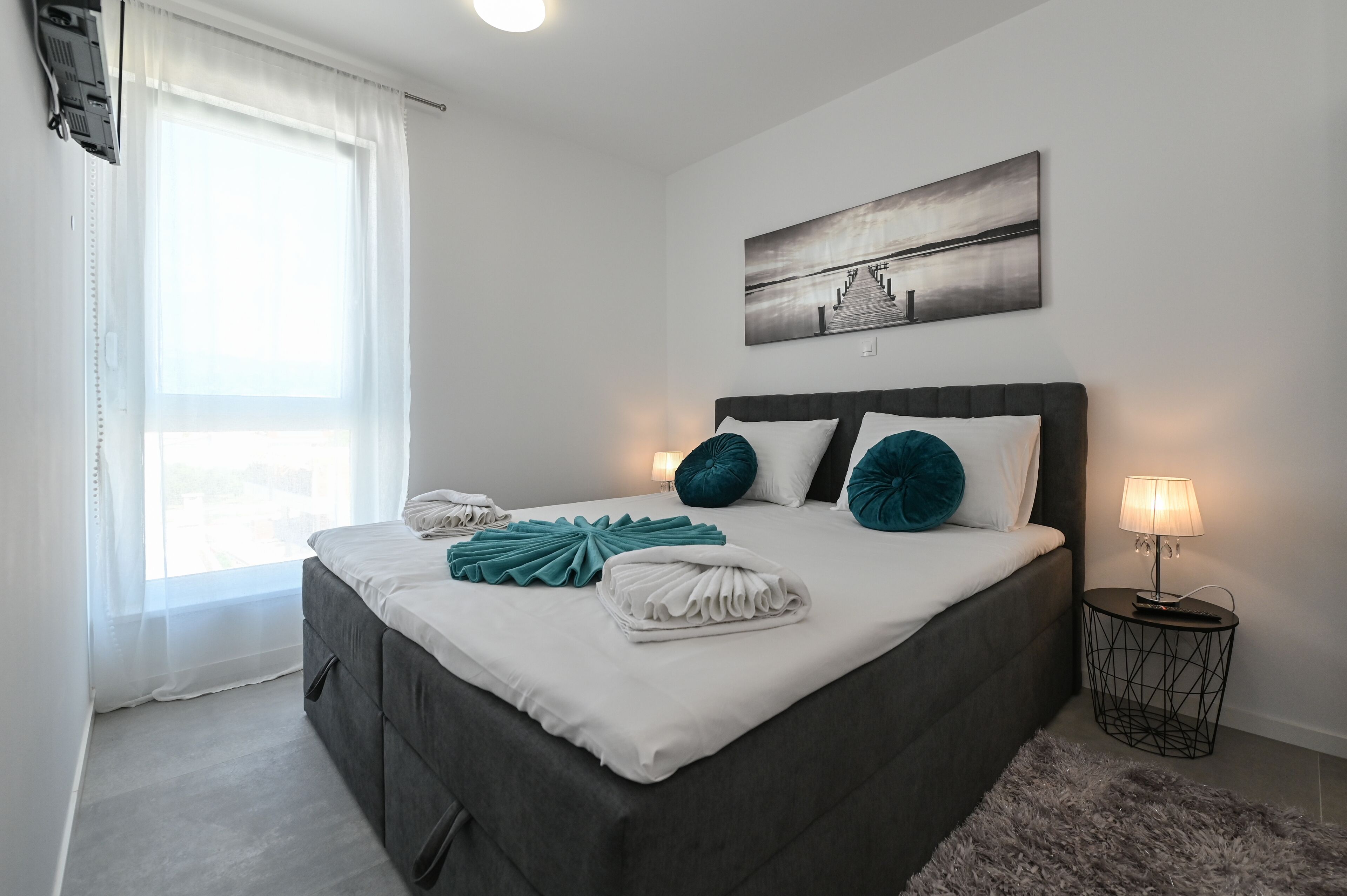 Una cama bien hecha con almohadas blancas y color turquesa, una mesita de noche con una lámpara, y un cuadro en la pared.