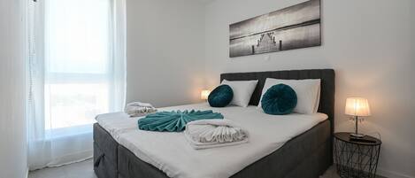 Una cama bien hecha con almohadas blancas y color turquesa, una mesita de noche con una lámpara, y un cuadro en la pared.