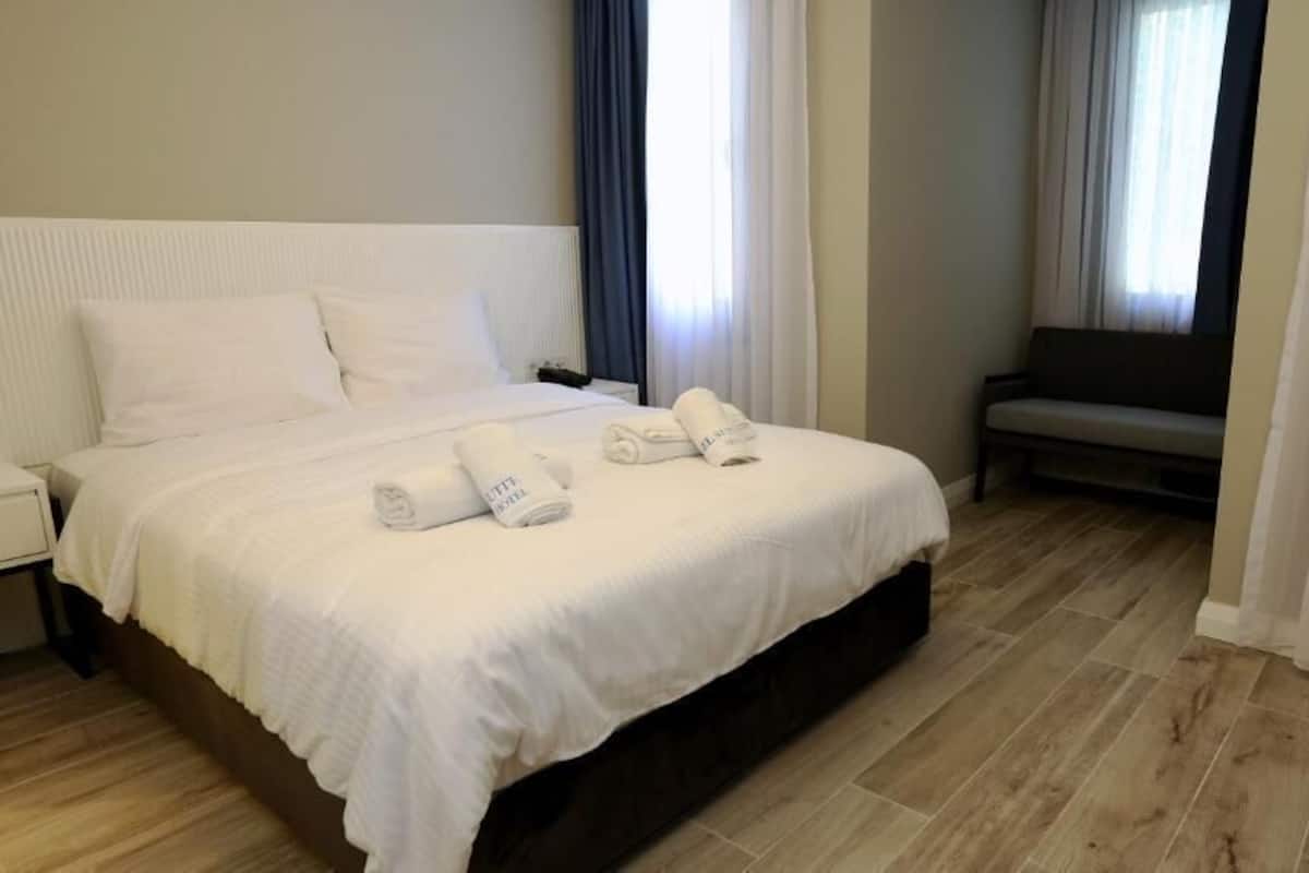 Una cama bien hecha con sábanas y toallas blancas, una mesita de noche y un banco en una habitación con suelo de madera y cortinas traslúcidas.
