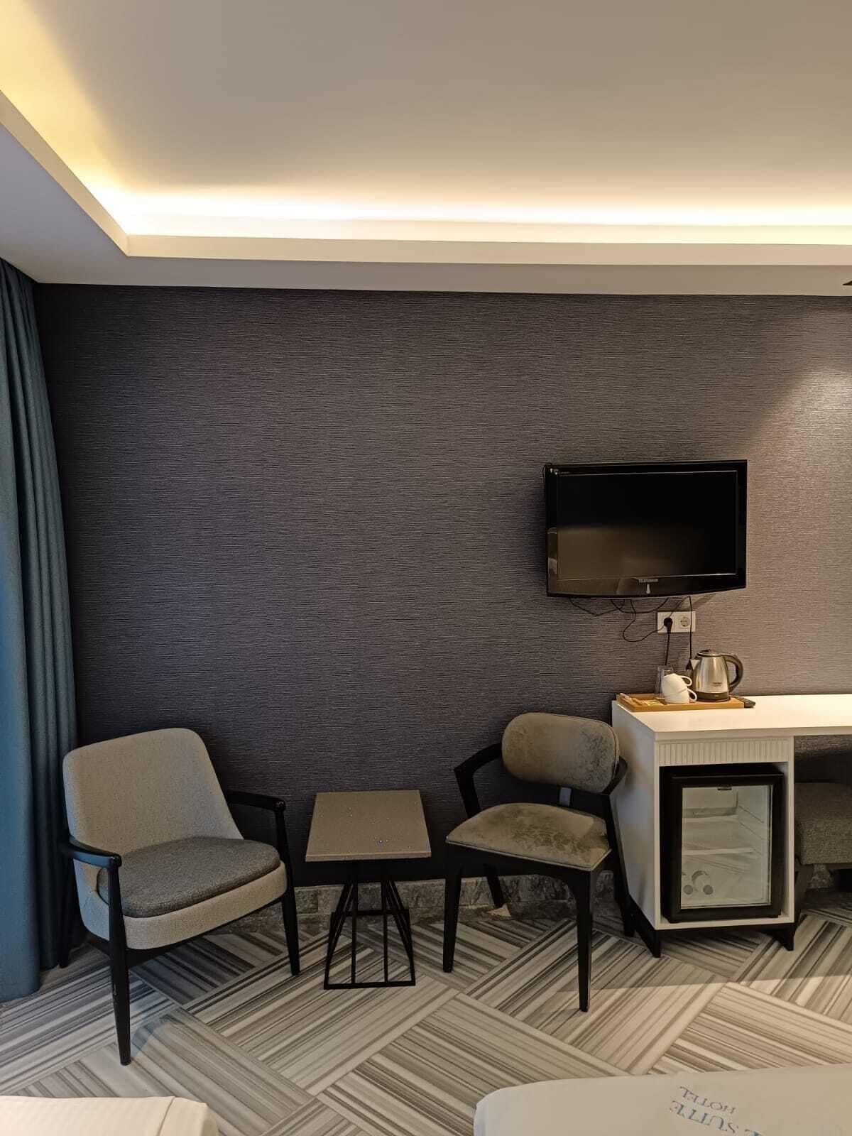 A hotel room with a dark textured wall, a flat-screen TV mounted above a desk, two chairs, a small table, and a minibar.