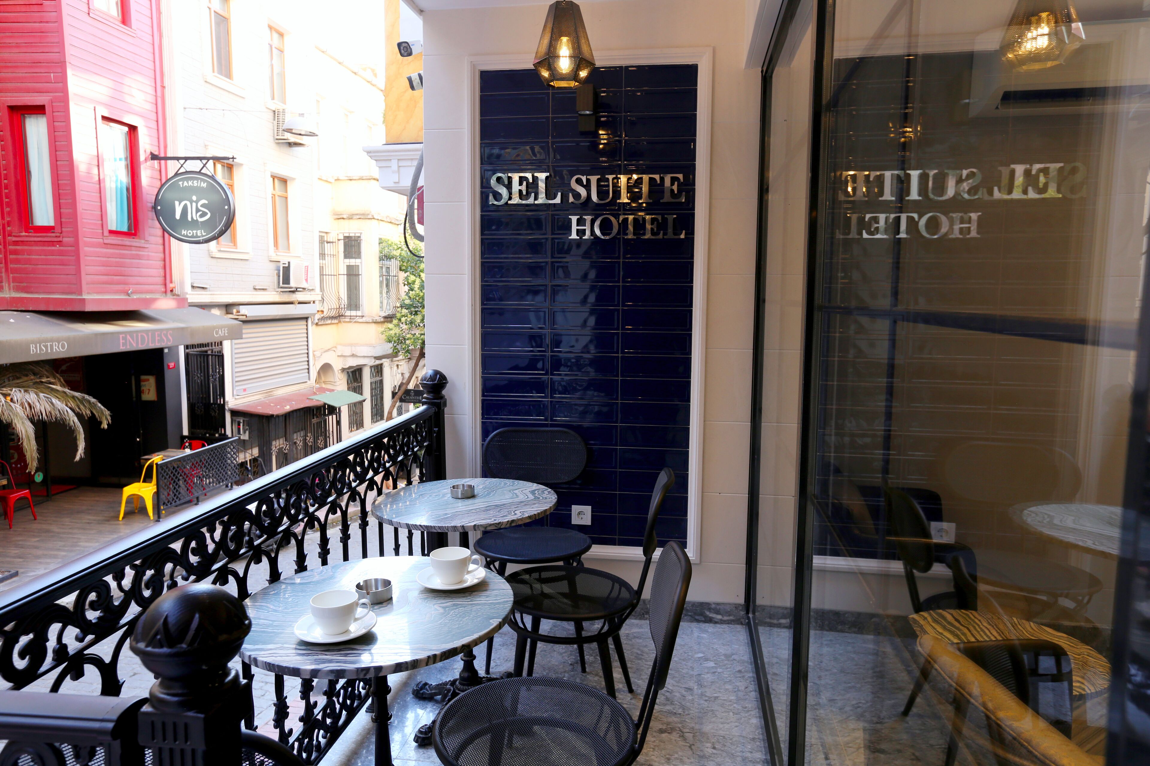 A hotel exterior with a blue sign reading "SEL SUITE HOTEL," outdoor seating with tables and chairs, and a visible entrance.