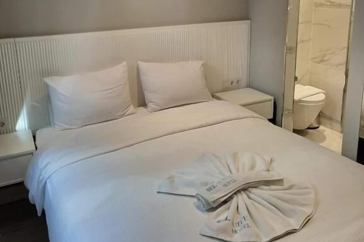 Habitación de hotel con una cama grande, ropa de cama blanca, una mesita de noche, un espejo y un baño visible a través de una puerta de cristal.