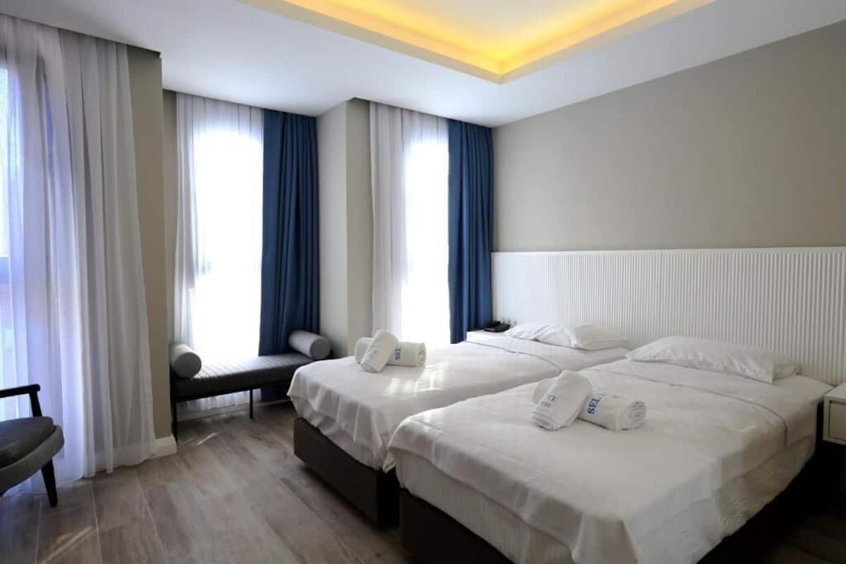 Una habitación de hotel con dos camas, un banco, una mesita de noche y una ventana con cortinas.