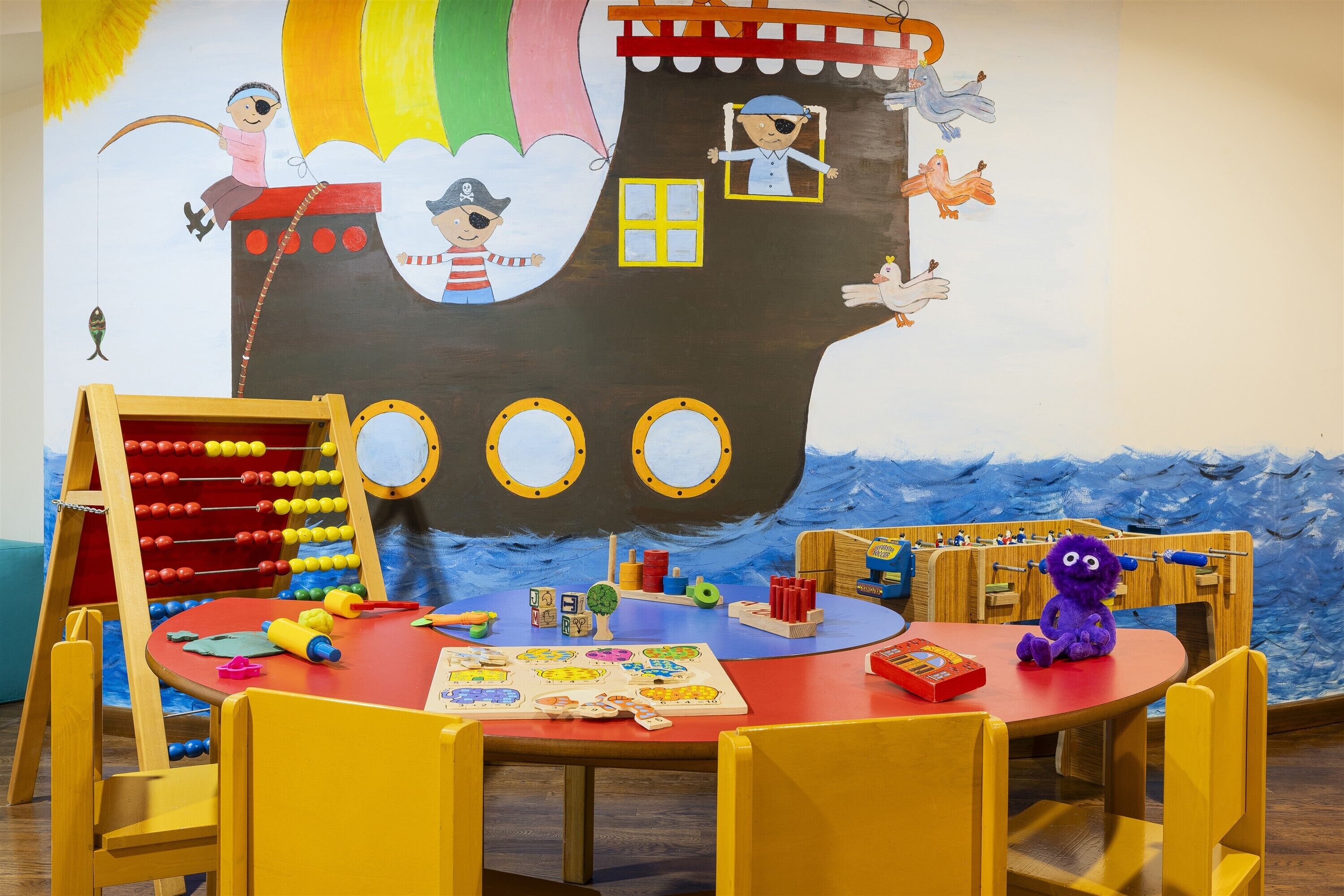 Un espace de jeux pour enfants agrémenté d’une grande fresque murale sur le thème des bateaux, de tables de jeux en bois et de divers jouets.