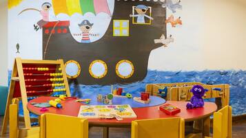 Un espace de jeux pour enfants agrémenté d’une grande fresque murale sur le thème des bateaux, de tables de jeux en bois et de divers jouets.