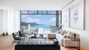 Ein modernes Wohnzimmer mit großem Fenster und Blick auf die malerische Küstenlandschaft, ein schwarzes Sofa, ein weißes Coucheck und ein Essbereich mit Stühlen.