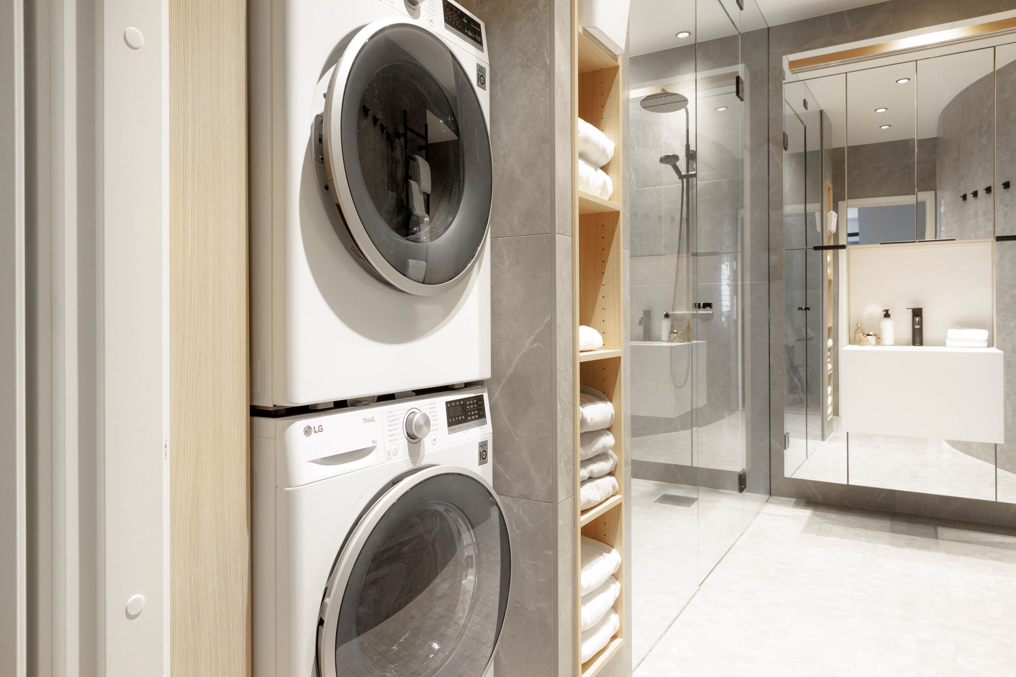A modern laundry room with stacked washing machines and dryers, a glass shower enclosure, and a minimalist bathroom with a white sink and toiletries.
