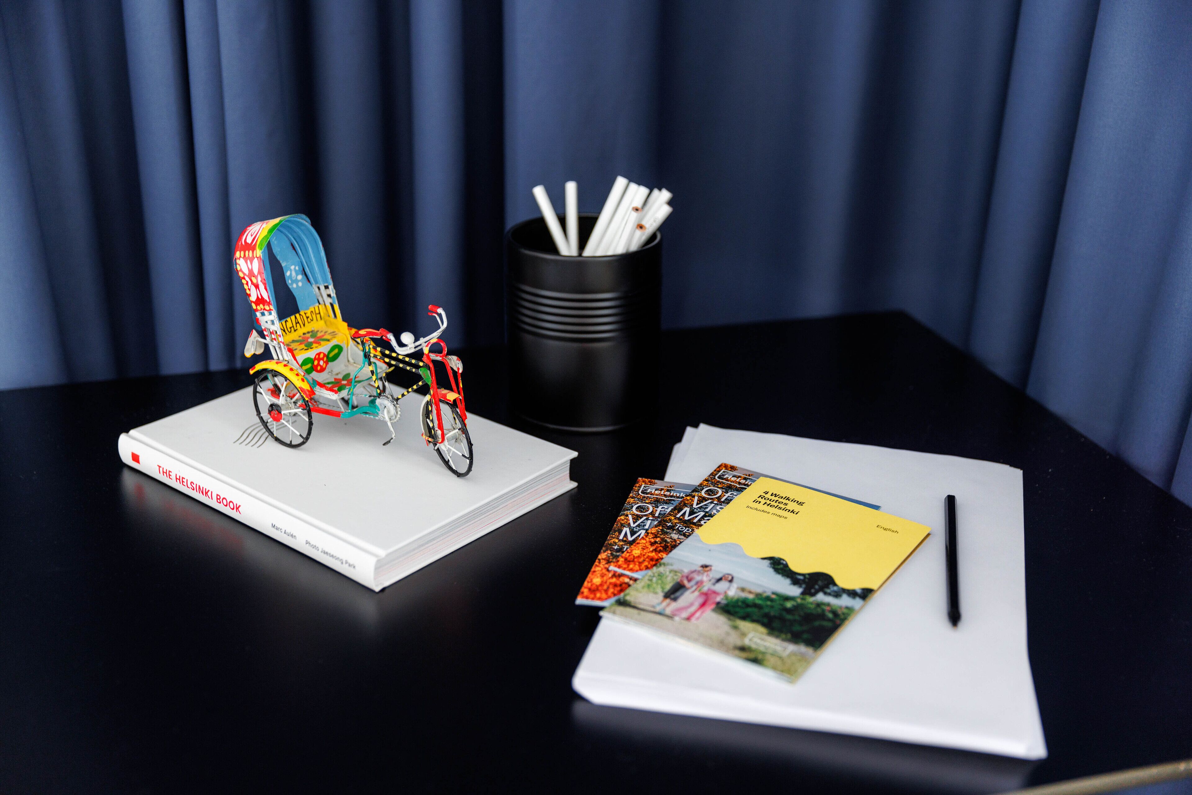 A table with a book titled "The Helsinki Book," a colorful bicycle, a black mug with straws, and a yellow envelope with a pen.