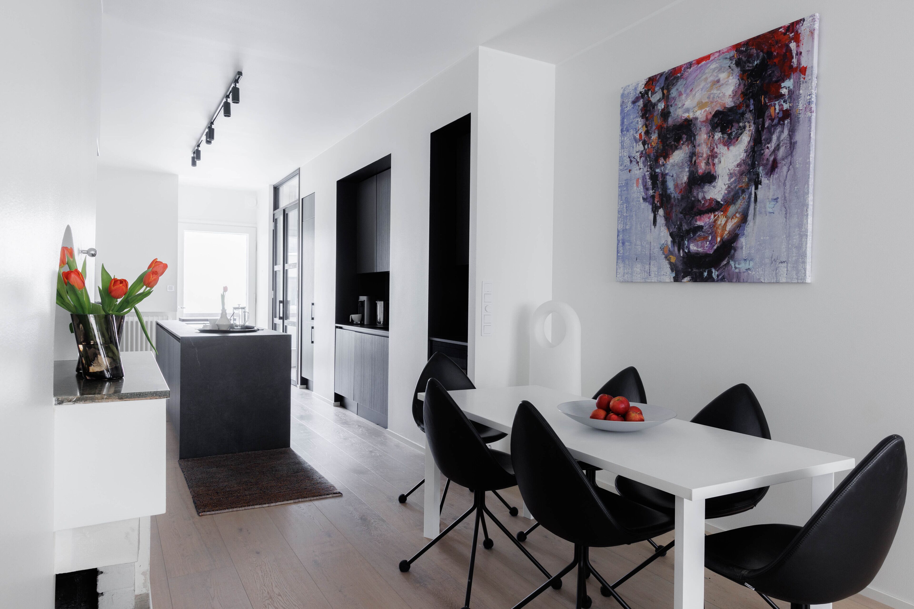 Une cuisine moderne avec un îlot central, des chaises noires disposées autour d’une table blanche et un grand tableau abstrait accroché au mur.