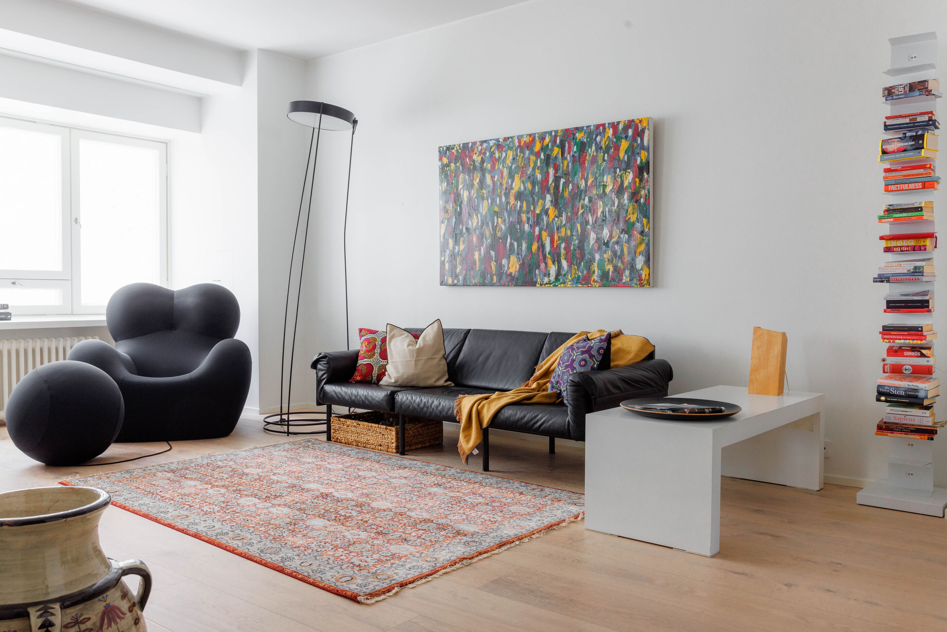 A modern living room with a black leather sofa, a colorful abstract painting, a bookshelf, and a patterned rug.