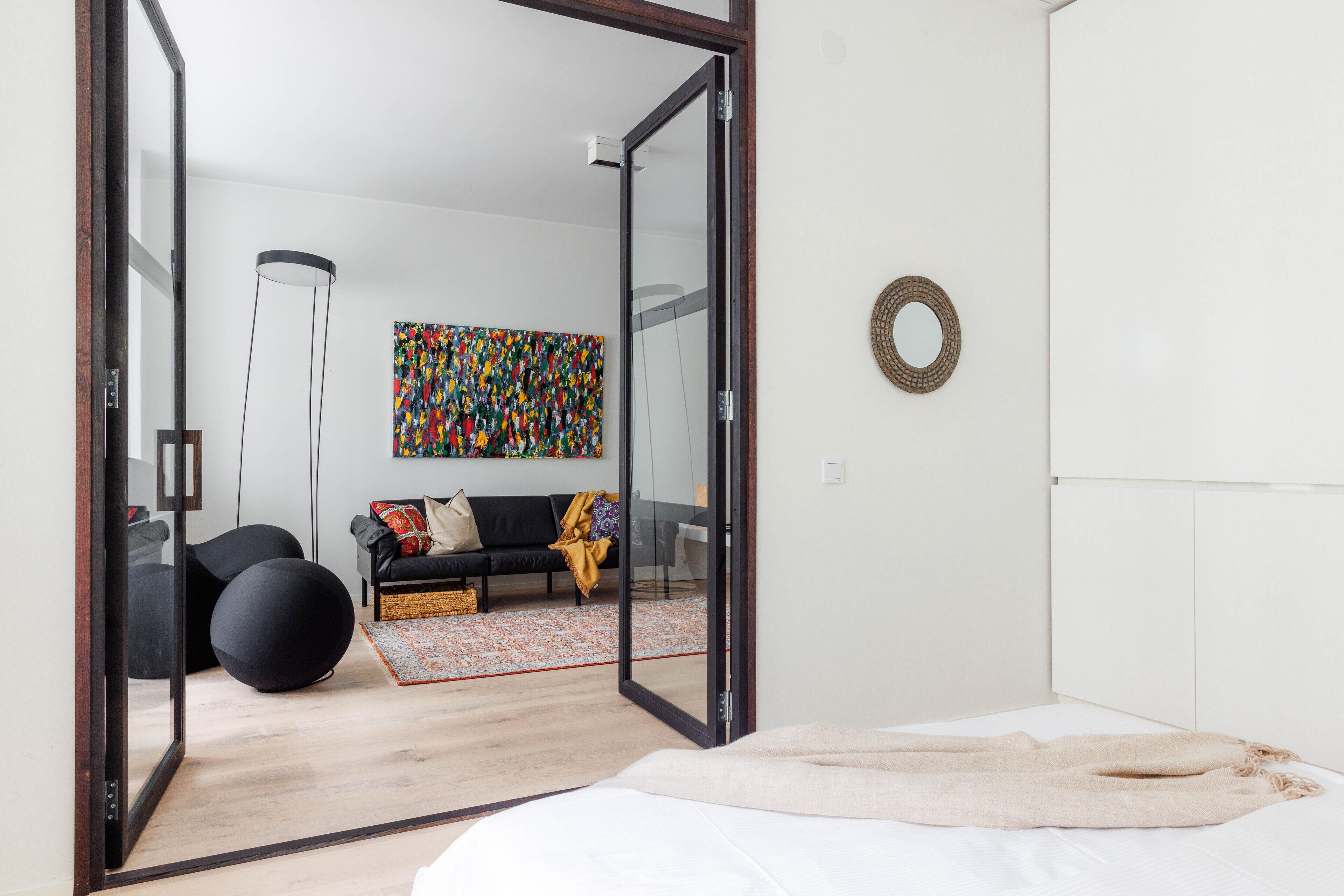 A modern bedroom with a large bed, a glass door leading to a living area, a colorful abstract painting, and a round mirror on the wall.