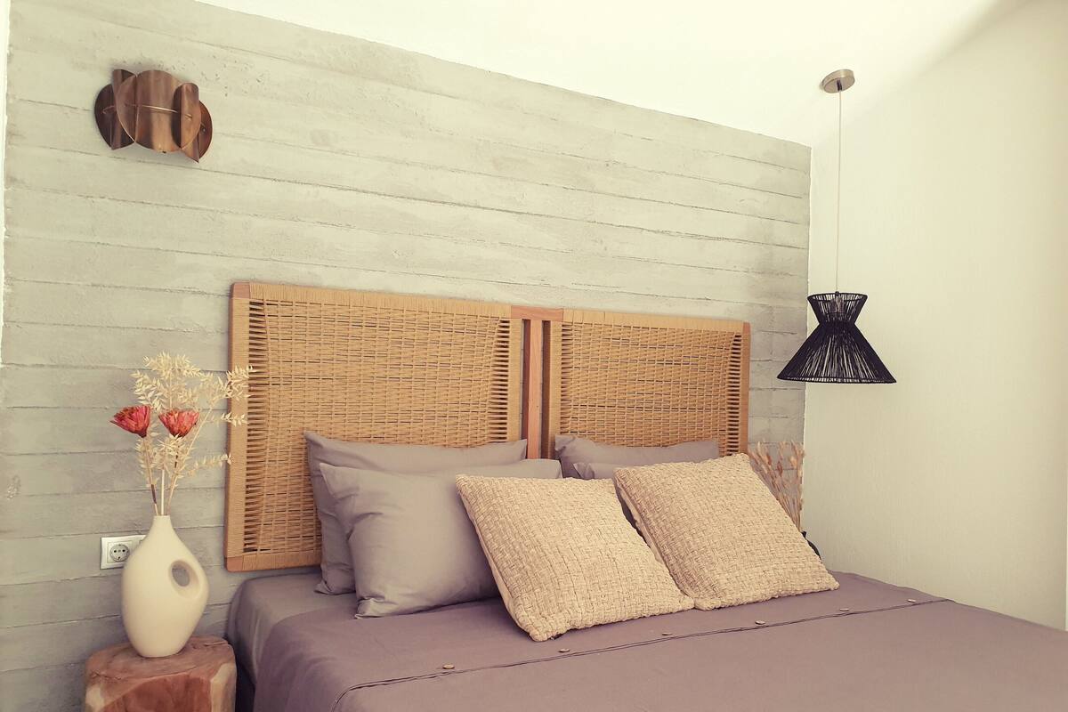 Uma cama com cabeceira de tecido, uma mesa de cabeceira com um vaso de flores secas e uma luminária pendente.