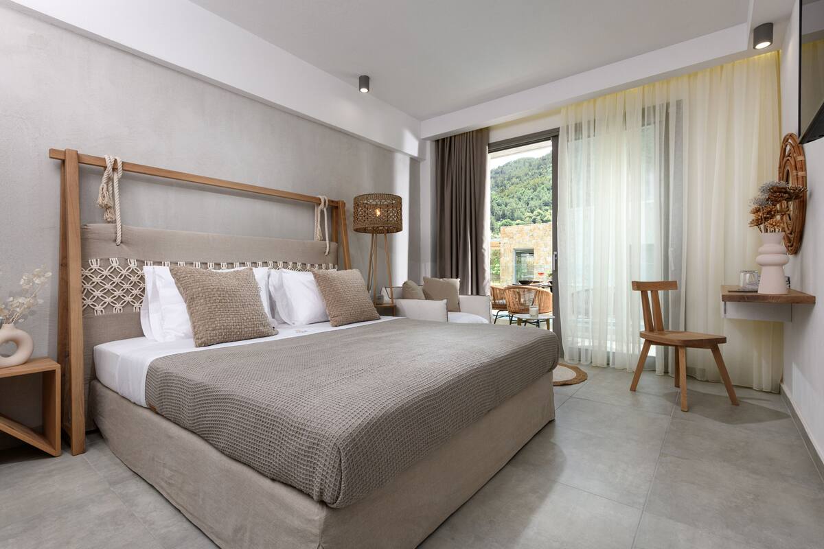 Quarto moderno com uma cama grande, cabeceira de madeira, mesa de cabeceira, uma cadeira e vista para o exterior.