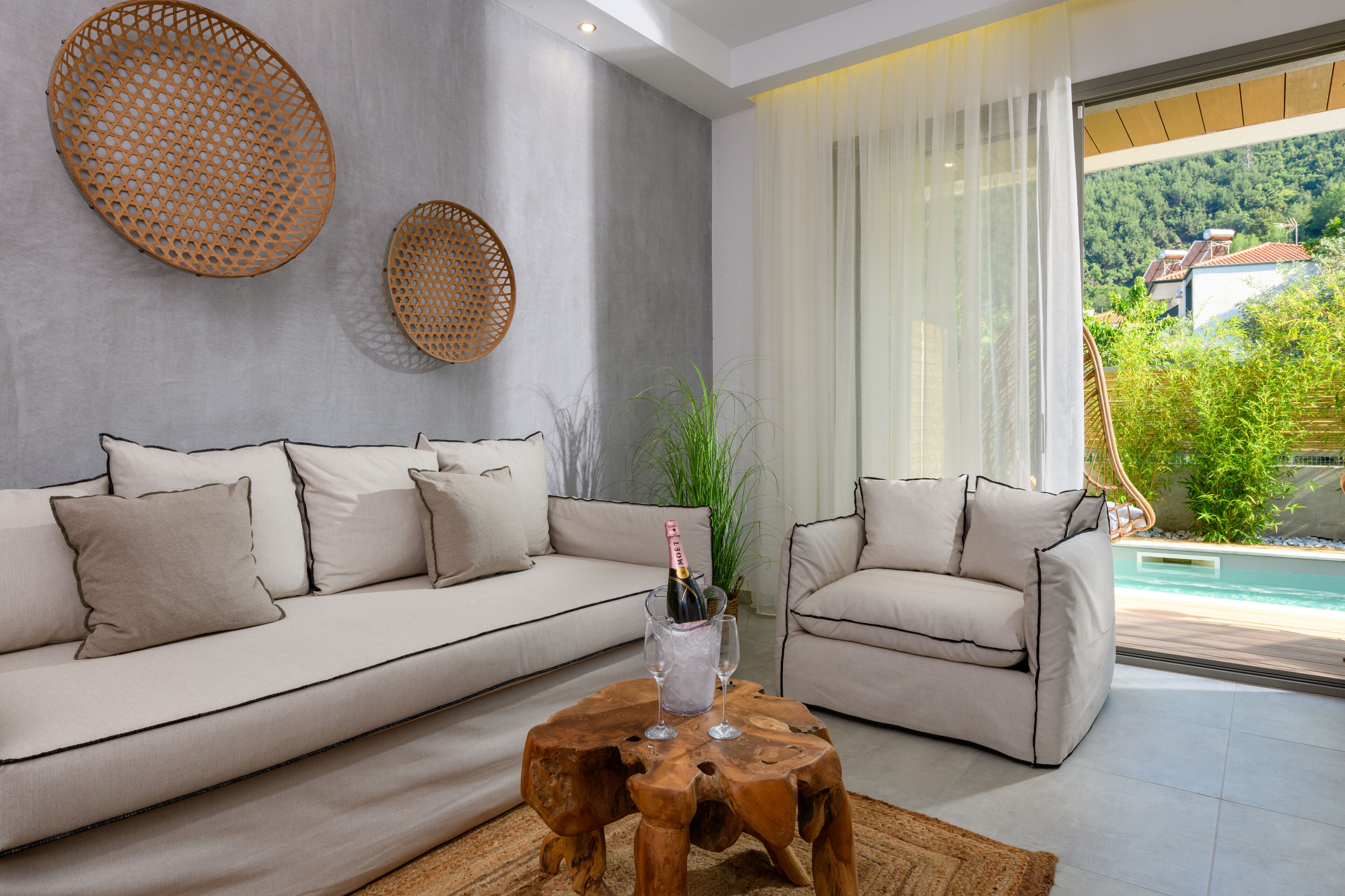 A modern living room with a beige sofa, a wooden coffee table with a bottle and glass, and a view of a pool and greenery outside.