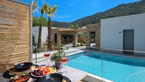 A modern outdoor pool area with a dining setup, including a table with fruits and drinks, surrounded by palm trees and a mountain backdrop. - Soho Exclusive Suites (Thasos)