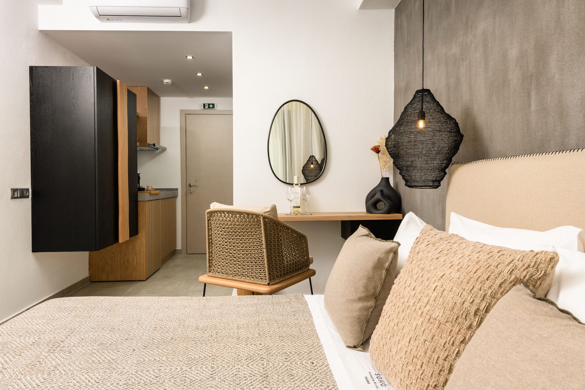 A modern hotel room with a bed, a sitting area with a chair, a small table, a mirror, and a hanging light fixture.