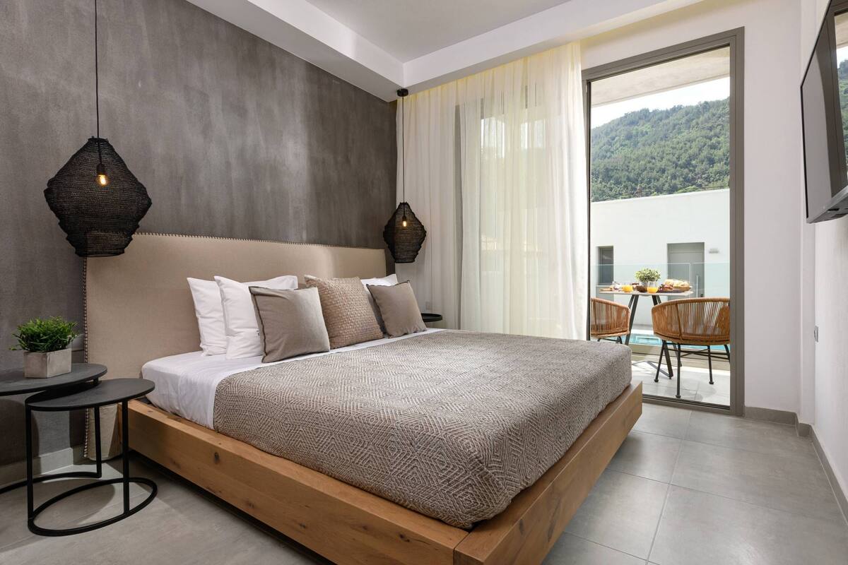 Quarto moderno com uma cama grande, uma mesinha de cabeceira e vista para uma varanda com cadeiras.