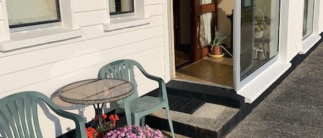 A white building with a window, a door, and a small table with chairs. There are potted plants and flowers outside.