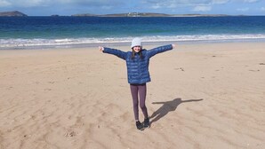 A person standing on a sandy beach with arms outstretched, facing a calm sea and distant hills under a clear sky.