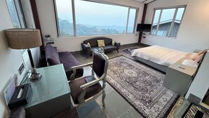 A modern hotel room with a large window, a sofa, a bed, a glass table, and a remote control on the bed. -  360 South, Khandala (Mawal)