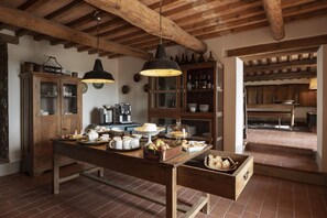 Una cocina rústica con vigas de madera, una gran mesa de madera con alimentos para el desayuno y un armario con puertas de vidrio.