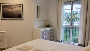 Ein Schlafzimmer mit einem Bett, einem Schminktisch, einem Spiegel und einem Fenster mit Blick auf Palmen.